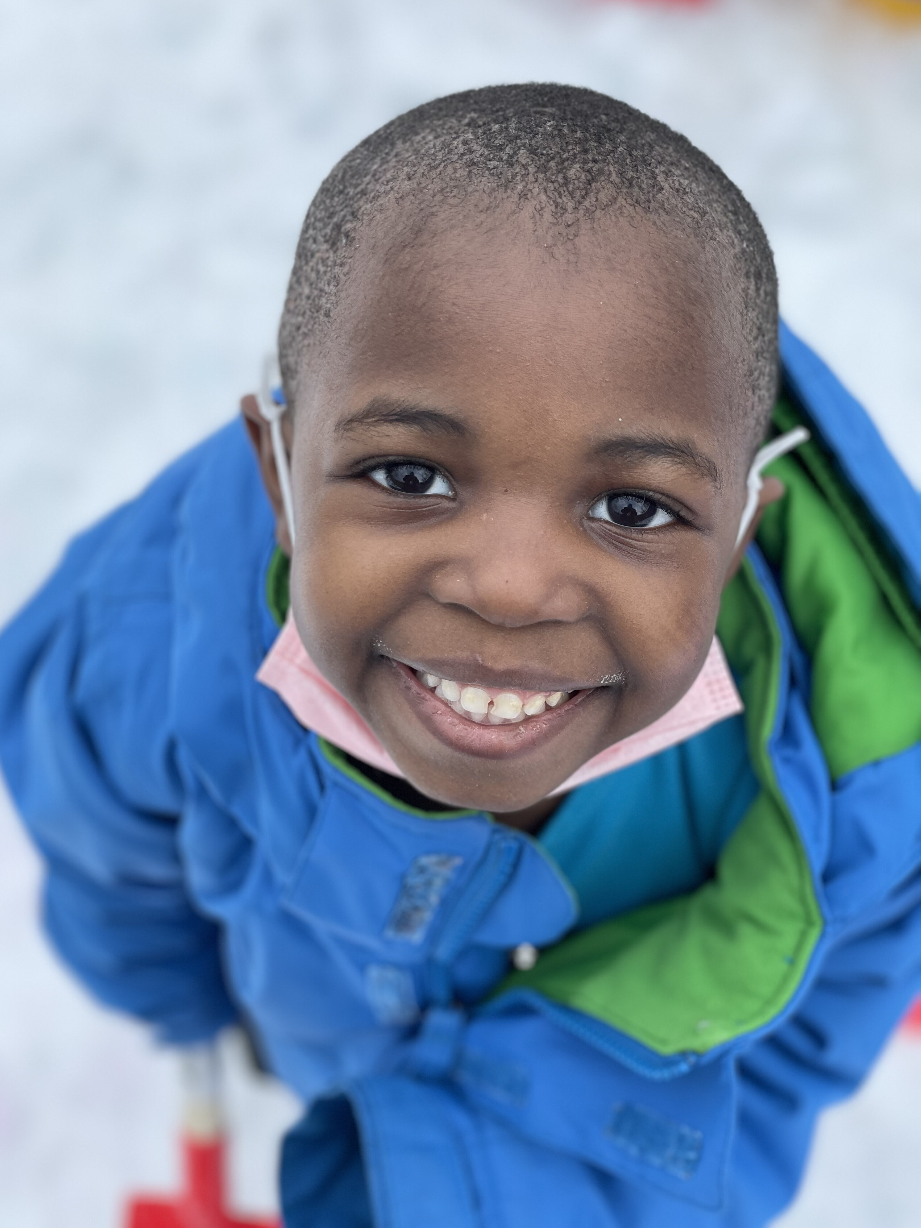 Photograph of a young black boy outside in Vermont smiling up towards the camera. The boy has short hair and is wearing a blue snow suit outside with snow around him.