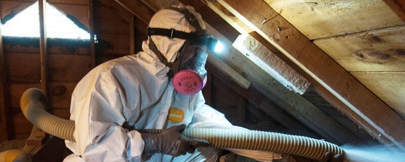 A crew member wears a respirator and tyvek suit as they insulate an attic with cellulose