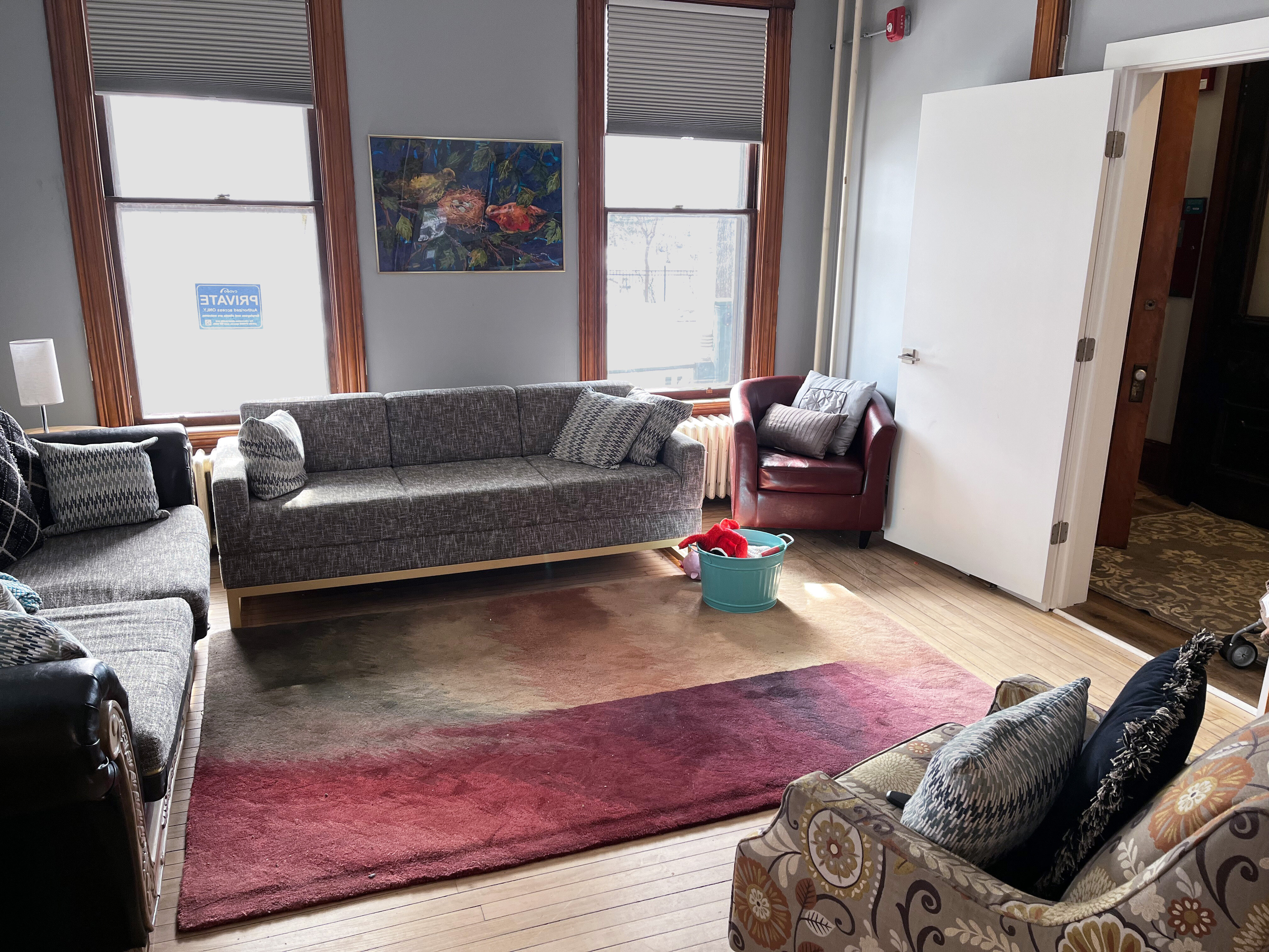 Bright sitting room with tall windows, gray walls, and wood floors. A large gray couch sits between two windows, with additional couches and chairs arranged around a large red and brown area rug. A burgundy leather armchair sits near an open white door, and a small blue bucket with red fabric rests on the floor in the sunlight. A framed painting hangs above the couch.